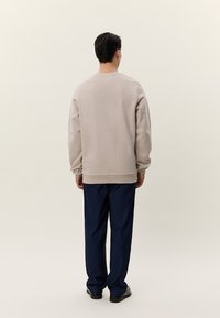 Muž stojaci chrbtom, oblečený v béžovej mikine, tmavomodrých nohaviciach a čiernych topánkach na svetlom jednoduchom pozadí.