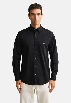 Férfi, aki fekete, testhezálló, hosszú ujjú, gombos inget és fehér nadrágot visel, egy egyszerű, világos háttér előtt áll.