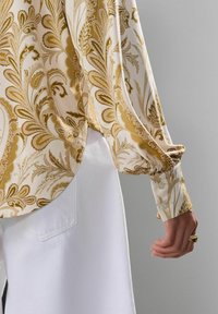 Blouse à manches longues en crème avec un motif floral doré, assortie à un tissu blanc. Présente des poignets à boutons et une texture lisse et brillante.