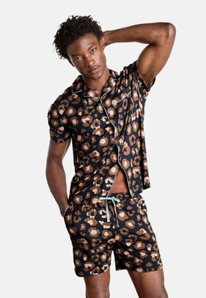 Man die een kort mouwen overhemd met knopen en korte broek met bruin en zwart abstract bloemmotief draagt, staand met één hand achter het hoofd en één in de zak.