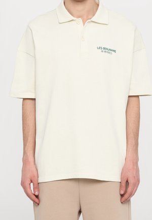 Cremfarbener Poloshirt mit kurzen Ärmeln, einem Kragen und einem grünen Logo auf der Brust. Aus weichem Material mit lockerer Passform.