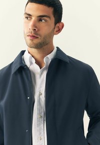 Giacca navy con finitura liscia, caratterizzata da un colletto a punta e chiusura con zip, indossata sopra una camicia bianca con bottoni visibili.