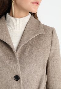 Beige wool-blend coat with a houndstooth pattern. Features a wide notched collar, a single black button, and a textured knit sweater underneath.