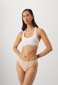 Valkoinen puuvillainen urheiluliivi, jossa on musta Calvin Klein -nauha ja beige saumatonta bikinit, sileä rakenne, muotoiltu malli ja minimalistinen tyyli.