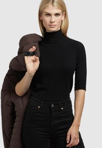 Zwarte coltrui met korte mouwen, gecombineerd met donkere jeans. Een bruine gewatteerde jas is over de schouder gedrapeerd. Zwarte armband om de pols.