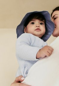 Lichtblauw gebreid babytruitje met lange mouwen, gecombineerd met een bijpassende blauwe bucket hat. Het kind wordt vastgehouden door een volwassene. Neutrale achtergrond.