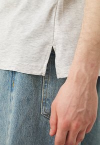 Šedé tričko s kulatým výstřihem a bočními rozparky, v kombinaci se světlými modrými džínami. Tkanina vypadá jemně s decentní texturou.