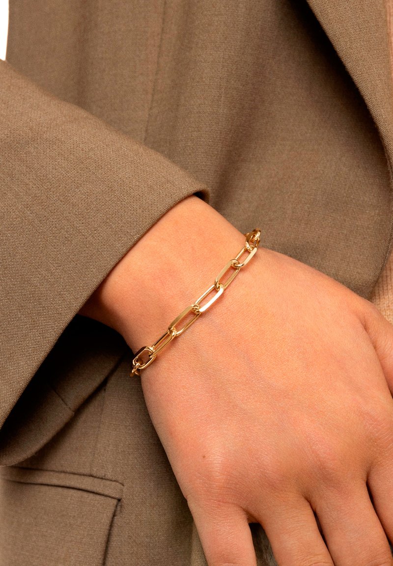 Pulsera de cadena de oro con eslabones ovalados, superficie lisa, llevada en una muñeca, combinada con una manga de tela neutra en un tono marrón apagado.