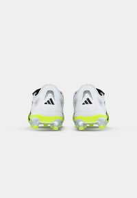 Une paire de chaussures de sport blanches avec des semelles jaunes fluo et un logo noir, vue de l'arrière sur un fond léger uni.