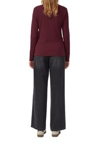Vrouw staat met de rug naar ons toe, draagt een bordeauxrode lange mouwen top, wijde vervaagde zwarte jeans en witte sneakers.