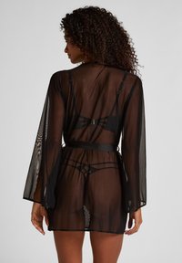 Γυναίκα με σγουρά μαλλιά που φοράει διαφανή μαύρη ρόμπα πάνω από ταιριαστό εσώρουχο, εμφανίζεται από πίσω μπροστά σε απλό γκρι φόντο.