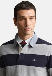 Pilko ir tamsiai mėlyno dryžuoto polo marškinėliai su šviesiai mėlynu apykakle, baltu sagų eiliu ir išsiuvinėtu logotipu ant krūtinės.