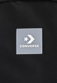 Černá textilní taška s obdélníkovou světle šedou gumovou nášivkou, na které je bílé logo hvězdy a šipky a tučný text "CONVERSE".