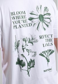 White cotton t-shirt featuring green floral graphics and text: “BLOOM WHERE YOU'RE PLANTED” and “RESPECT THE LOCALS.” Casual design.