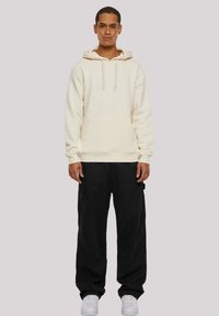 Sweat à capuche en polaire crème avec une poche avant, capuche à cordon, associé à un pantalon noir ample et des baskets blanches, se tenant devant un fond gris.