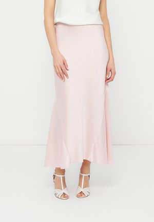 Žena má na sebe svetloružovú sukňu dĺžky po členky s jemnou textúrou, ktorú dopĺňajú biele sandále na podpätku a biely tielkový top.