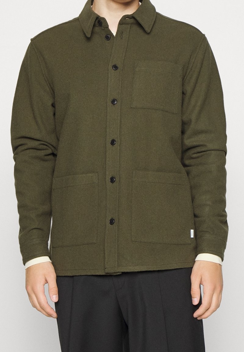 Homme portant une veste boutonnée vert olive avec trois poches frontales par-dessus une chemise légère à manches longues et un pantalon noir.