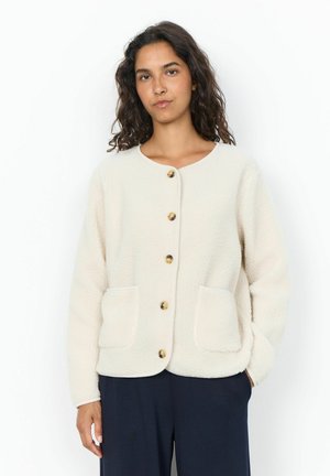 Donna con capelli ricci scuri indossa una giacca in pile bianca con bottoni e due tasche frontali e pantaloni scuri, in piedi davanti a uno sfondo bianco.