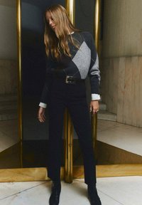 Maglione argyle grigio e nero con polsini bianchi, abbinato a pantaloni neri a vita alta e stivali alla caviglia. Dettaglio della fibbia della cintura rettangolare argentata.