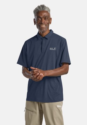 Älterer Mann mit kurzen grauen Haaren trägt ein marineblaues Jack Wolfskin Polohemd und beige Hose, lächelt und hält die Hände sanft verschränkt.