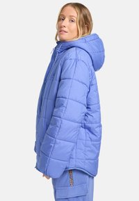 Lysblå polstret jakke med hætte, der har lynlås lukning, en boxy form og subtile søm mønstre. Der er detaljer som snore.