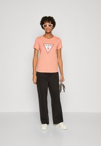 Ferskenfarvet T-shirt med korte ærmer og et sort, trekantet "GUESS" logo. Kombineret med sorte, vide bukser og hvide sneakers. Model holder en sølvfarvet taske.