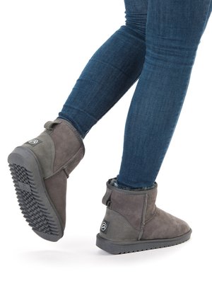 Person iført blå jeans og grå ankelstøvler med gummisåler, som går med det ene ben løftet mod en hvid baggrund.