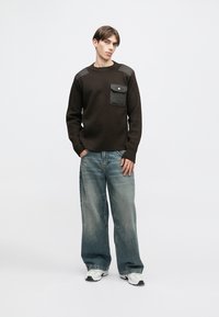 Brūns džemperis ar plecu uzlikām, priekšējā kabatā, un pelēkām detaļām, kombinēts ar gaiši zilām brīva piegriezuma džinsām un baltām sporta kurpēm.