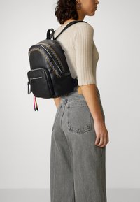 Schwarzer Leder-Rucksack mit dekorativer, mehrfarbiger Stickerei, vorderer Reißverschlusstasche und Quast-Details. Getragen über grauen, weit geschnittenen Jeans.