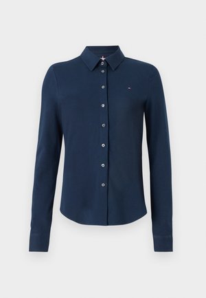 Chemise à manches longues bleu marine avec col, présentant un devant boutonné et un petit logo brodé sur la poitrine. Texture de tissu lisse.
