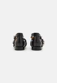 Zapatos de charol negros con una correa trasera y hebilla de acento plateado. Presentan un tacón bajo y una superficie lisa y brillante.