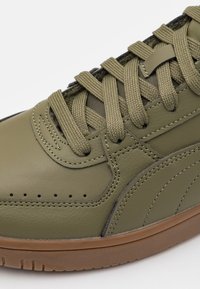 Detailní záběr na olivově zelené kožené tenisky s tlustými odpovídajícími tkaničkami a gumovou podrážkou v barvě gumy na bílém pozadí.