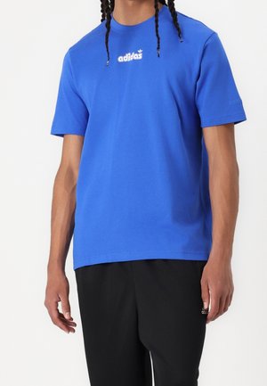 Personne portant un t-shirt bleu Adidas avec un logo blanc et un pantalon Adidas noir, debout devant un fond uni.