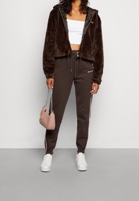 Brązowa kurtka z sztucznego futra na zamek, krótka, nałożona na białą bluzkę crop top, w zestawieniu z brązowymi dresami z białymi paskami po bokach i białymi sneakersami. Różowa torebka.