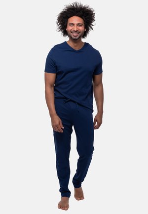 Uomo con i capelli ricci che sorride, indossa una camicia a maniche corte blu scuro e pantaloni abbinati, a piedi nudi, cammina in avanti su uno sfondo semplice.