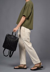 Chemise olive à manches courtes, pantalon beige ample, sac à dos noir texturé, sandales plates marron. Design simple et pratique avec des couleurs neutres.