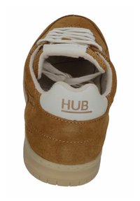 Braune Wildledersneaker mit weißen Akzenten, strukturiertem Obermaterial, flachen Schnürsenkeln und einem markierten Fersenbereich. Die Sohle ist aus cremefarbenem Gummi.