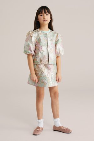 Ragazza giovane in piedi che indossa una giacca in broccato floreale pastello e una gonna abbinata, calzini bianchi e ballerine rosa.