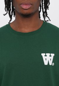 Green cotton t-shirt with a ribbed crew neckline and a white stylized "W" logo on the left chest. Smooth texture, standard fit.