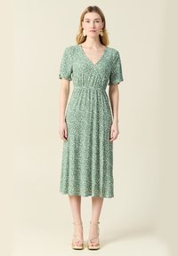 Kwiatowa zielona sukienka midi z krótkimi rękawami, dopasowanym gorsetem i rozkloszowanym dołem. Posiada dekolt w kształcie litery V oraz subtelny wzór w białe kwiaty. Złote sandały na obcasie.