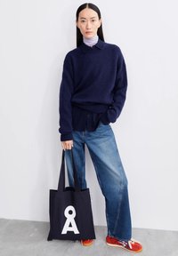 Femme portant un pull bleu marine sur une chemise à col et un jean bleu, tenant un sac fourre-tout noir avec la lettre blanche Å, debout devant un mur blanc.