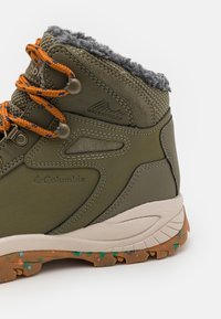 Bota impermeable en verde olivo con forro de piel sintética gris, cordones naranjas y suela marrón texturizada con manchas de colores.