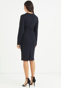 Langärmliges, tailliertes dunkelblaues Kleid mit Reißverschluss hinten, knielanger Schlitze und glattem Stoff. Kombiniert mit schwarzen Ankle-Strap-High Heels.