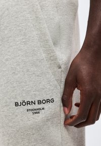 Κοντινό πλάνο σκουρόχρωμου χεριού που ακουμπά κοντά στην τσέπη ανοιχτόχρωμου γκρι φόρμας με τυπωμένο "BJÖRN BORG STOCKHOLM 1956".