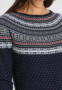Marineblaues Sweater mit einem strukturierten Muster, der weiße und rote geometrische Designs über die Brust und Schultern aufweist, Rundhalsausschnitt und lange Ärmel.