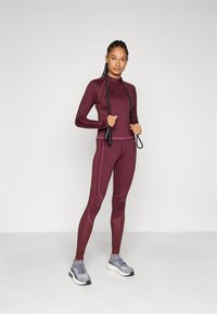 Mørkerød treningssett bestående av en langermet topp med høyt krage og matchende leggings, aksentuert med rosa sømmer og et treningshoppetau.