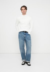 Cămașă albă cu mâneci lungi, blugi albaștri din denim, curea neagră și pantofi negri cu accente albe. Modelul stă drept, cu un fundal simplu.