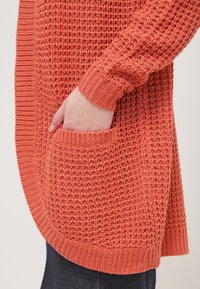Cardigan lavorato a maglia in arancione corallo con un motivo testurizzato, polsini a coste e una tasca frontale. Il design con spacco laterale offre una vestibilità rilassata.