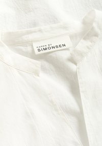 Blouse blanche avec un col en V et un col court. Fabriquée en tissu léger, avec une étiquette tissée portant "KAREN BY SIMONSEN" en noir.