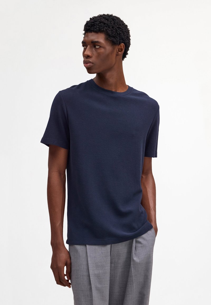 Mand iført en marineblå kortærmet skjorte og grå plisserede bukser, der ser til venstre med en hånd i lommen.